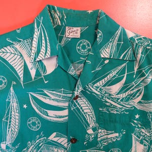 May include: Teal button-up shirt with a white sailboat pattern. The shirt features a classic collar, a chest pocket, and brown buttons. The tag reads "Shakees's by Honolulu".