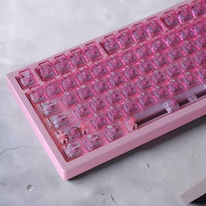 May include: A mechanical keyboard featuring a solid pink frame and translucent pink keycaps. The keys display white lettering, including standard QWERTY layout characters and specific labels such as Tabulator, Caps Lock, Shift, Control, and Super.