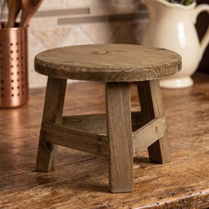 Könnte beinhalten: Ein kleiner, runder Holzhocker mit natürlichem braunem Finish. Der Hocker verfügt über eine kreisförmige Oberfläche, die von vier stabilen, geraden Beinen getragen wird, welche durch horizontale Holzstreben nahe der Basis verbunden sind.
