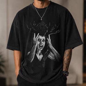 Puede incluir: Una camiseta negra de manga corta con un estampado gráfico de una persona con la boca abierta y las manos levantadas hacia la cabeza, enmarcada por formas oscuras similares a astas. La camiseta tiene un aspecto desteñido y cuello redondo.