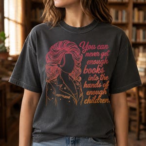 Puede incluir: Una camiseta de manga corta gris carbón con una ilustración de líneas rosas y naranjas de una persona con cabello voluminoso y una chaqueta con cuello. El texto dice, You can never get enough books into the hands of enough children.