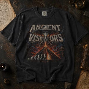 May include: A charcoal gray t-shirt featuring a graphic print of a large alien spacecraft hovering over a pyramid. The text reads ANCIENT VISITORS and THE ASCENT BEGINS. Below the craft, a human evolution sequence leads toward the pyramid structure.