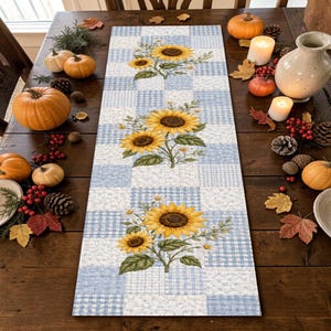May include: A rectangular table runner featuring a patchwork pattern of light blue and white gingham and floral squares. The runner is decorated with three distinct clusters of yellow sunflowers and green leaves arranged along the center line.