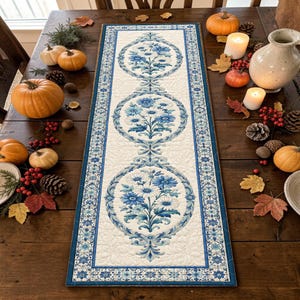 Puede incluir: Un camino de mesa acolchado rectangular con fondo blanco y tres medallones florales azules repetidos. El diseño incluye bordes intrincados con volutas azules y una textura cosida detallada en toda la superficie de la tela.