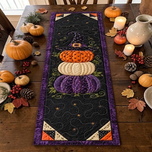 May include: A rectangular quilted table runner featuring a central stack of three pumpkins in orange, white, and purple, topped with a witch hat. The black fabric background is decorated with embroidered stars and vines, framed by a purple border.