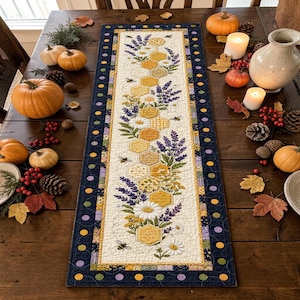 Puede incluir: Un camino de mesa acolchado rectangular que presenta un panel central de color crema con un patrón repetitivo de panales amarillos, ramas de lavanda moradas, margaritas blancas y pequeñas abejas, bordeado por una tela azul oscuro con puntos amarillos.