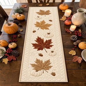 May include: A rectangular quilted table runner in cream fabric featuring embroidered autumn motifs including brown maple leaves, white pumpkins, and small acorns, with a diamond-stitched border pattern running along the entire outer edge.