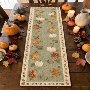 May include: A rectangular quilted table runner featuring a sage green background with a repeating pattern of white pumpkins, orange and brown autumn leaves, and acorns. The runner has a cream-colored border decorated with small fall leaves.