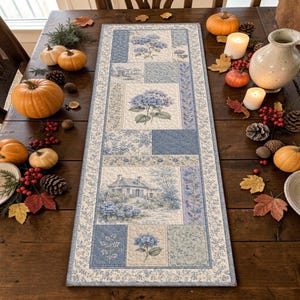 May include: A rectangular quilted table runner featuring a patchwork design with blue and white floral patterns, hydrangea illustrations, and a landscape scene of a house, bordered by a thin blue floral print on a cream-colored background.