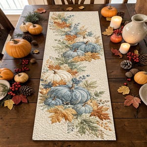 May include: A rectangular table runner featuring a printed pattern of blue, white, and orange pumpkins surrounded by autumn leaves and berries. The fabric has a textured, quilted appearance with a cream-colored background and decorative stitching.