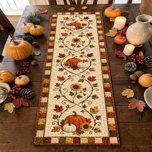 May include: A rectangular quilted table runner featuring a cream-colored center panel with an intricate pattern of orange pumpkins, yellow sunflowers, and red and green autumn leaves, framed by a border of small multicolored fabric squares.