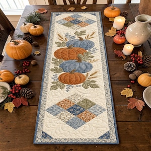 Puede incluir: Un camino de mesa rectangular de tela con un diseño central vertical de cuatro calabazas apiladas en tonos naranja y azul, rodeadas de hojas y tallos de trigo, con extremos acolchados de patrones de diamantes en estampados florales.