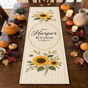 May include: A rectangular quilted table runner featuring a cream-colored fabric with an intricate sunflower and wheat pattern. The center displays a decorative gold-bordered frame with the text The Harper Kitchen printed in a black script font.