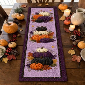May include: A rectangular quilted table runner featuring a light purple background with a subtle spiderweb pattern. The runner displays three vertical stacks of pumpkins in orange, white, purple, and black, accented with colorful autumn leaves.