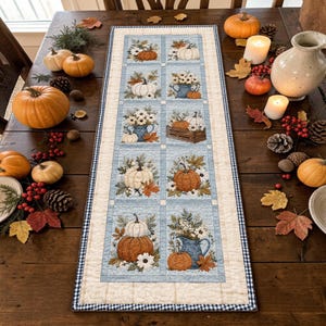 Puede incluir: Un camino de mesa acolchado rectangular con una cuadrícula de paneles que muestran ilustraciones de calabazas, hojas de otoño y arreglos florales en jarras azules. El camino tiene un borde color crema con un acabado de cuadros azules y blancos.