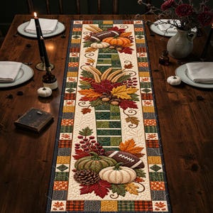 Puede incluir: Un camino de mesa acolchado rectangular con un diseño central de hojas de otoño, calabazas, espigas de trigo, bellotas y balones de fútbol americano. El borde presenta un patrón de patchwork en tonos naranja, verde, amarillo y marrón.