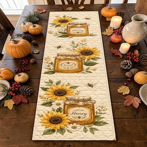 May include: A rectangular table runner featuring a repeating pattern of honey jars and sunflowers on a cream-colored background with a quilted honeycomb texture. The jars are labeled with the text Pure Honey, Sweet as can bee, and Local Honey.