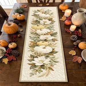 May include: A rectangular quilted table runner featuring a printed pattern of white pumpkins, cream-colored magnolia flowers, green foliage, and wheat stalks on a cream background, displayed on a dark wooden table with scattered pumpkins.