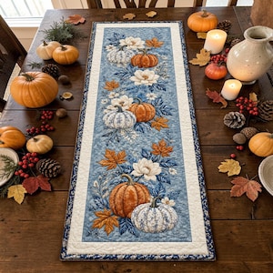 May include: A rectangular fabric table runner featuring a blue background with a repeating pattern of orange pumpkins, white pumpkins, white magnolia flowers, and autumn leaves in shades of orange and brown, finished with a white and blue border.