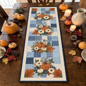 Puede incluir: Un camino de mesa acolchado rectangular con un patrón de cuadros azules y blancos, decorado con motivos repetidos de calabazas naranjas, flores blancas y hojas de otoño. El camino tiene un borde azul oscuro sobre una mesa de madera.