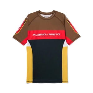 May include: A short-sleeve rash guard featuring a color-blocked design in brown, red, black, and yellow. The chest displays the white text ALBINO & PRETO COMPETITION TEAM. The sleeves feature red and white accents with the brand name printed on them.