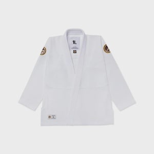 May include: A white martial arts gi jacket featuring long sleeves and a structured collar. The garment includes a small rectangular label on the lower front with the text 1L, and circular brown and gold patches on each shoulder area.