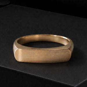 May include: A solid gold-toned ring featuring a flat, rectangular bar top section. The band has a smooth, matte finish with a consistent metallic colour throughout, showcasing a clean, unadorned design with a rounded inner profile for comfort.