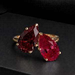 Peut inclure: Une bague en or ornée de deux pierres précieuses rouge foncé en forme de poire, placées côte à côte. Les pierres sont facettées et maintenues par de petites griffes en or, formant un design à deux pierres sur un anneau en or poli.
