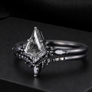 May include: A black metal ring set featuring a kite-shaped rutilated quartz centre stone. The band is accented with small black stones in a chevron-shaped arrangement, creating a layered appearance with a dark, polished finish throughout the design.