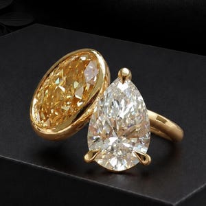 Peut inclure: Une bague ouverte en or jaune ornée de deux pierres distinctes. L'une est une grande gemme jaune de taille ovale sertie clos, tandis que l'autre est une gemme blanche transparente de taille poire fixée par des griffes, formant un duo.