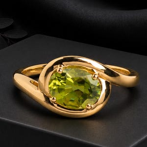 May include: A gold ring featuring a central oval-cut lime green gemstone held by four prongs. The band has a smooth, polished finish and curves around the stone in a bypass style, creating a continuous, fluid shape that encircles the setting.