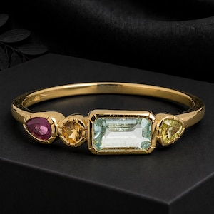 May include: A gold ring featuring a central emerald-cut light blue gemstone flanked by a small round yellow stone and a pear-shaped pink stone on one side, and a triangular yellow stone on the other side, set on a thin polished gold band.