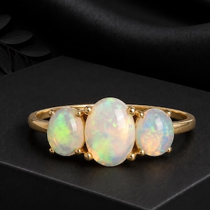 Puede incluir: Un anillo de oro con tres ópalos de forma ovalada dispuestos en fila. El ópalo central es más grande que las dos piedras laterales, y cada gema muestra destellos iridiscentes de color verde, naranja y amarillo sobre una base blanca lechosa.