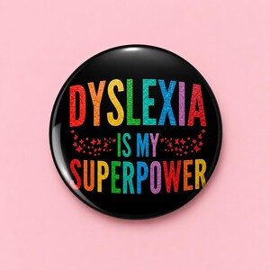 May include: A round black pin-back button featuring the multi-coloured glittery text DYSLEXIA IS MY SUPERPOWER in a bold sans-serif font. The design includes small red star accents scattered below the word DYSLEXIA on a dark background.