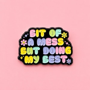 May include: A black enamel pin with a wavy, cloud-like border featuring the text BIT OF A MESS BUT DOING MY BEST in puffy, multi-coloured pastel letters. The design includes small decorative flower and star icons in pink, yellow, and blue.