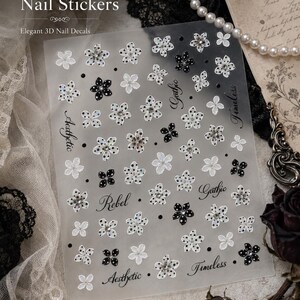 May include: A rectangular sheet of nail stickers featuring a variety of small five-petalled flowers in black, white, and iridescent finishes. The sheet includes text elements reading Gothic, Aesthetic, Rebel, and Timeless in a cursive script font.