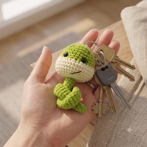 May include: A crochet snake keyring featuring a green and cream-coloured yarn body with black bead eyes and a stitched smile, attached to a silver metal ring holding a collection of various brass and grey metal keys for everyday use.