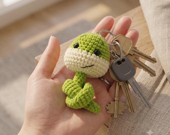 May include: A crochet snake keychain featuring a green and cream-colored yarn body with black bead eyes and a stitched smile, attached to a silver metal keyring holding a collection of various brass and grey metal keys for everyday use.