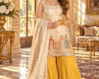 May include: A cream-colored sleeveless tunic with intricate multi-colored embroidery at the hem, paired with wide-leg mustard yellow trousers and a matching cream-colored shawl featuring a decorative geometric border pattern.