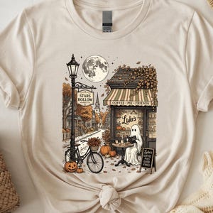 May include: A cream-colored t-shirt featuring a graphic illustration of a town scene with a ghost sitting at a table outside a shop labeled Luke's. The design includes a street lamp, a bicycle, pumpkins, a full moon, and text reading Stars Hollow.