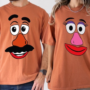 May include: Two rust-colored short-sleeve t-shirts. The left shirt features a graphic of a face with black eyebrows, a large orange nose, and a black mustache. The right shirt features a face with purple eyeshadow, long eyelashes, a pink nose, and red lips.