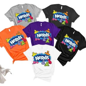May include: A collection of six short-sleeve t-shirts in gray, black, orange, purple, and white. Each shirt features the blue NERDS candy logo surrounded by colorful, small, round character illustrations in yellow, orange, pink, purple, and green.