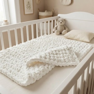Op de afbeelding: Een witte grofgebreide deken met een dikke, zachte textuur die over een wit houten ledikantmatras ligt. De deken heeft een opvallend patroon van lussen en ligt naast een lichtbeige kussen en een kleine lichtbruine teddybeer.