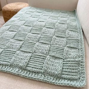 Op de afbeelding: Een licht saliegroene gehaakte deken met een mandvlechtpatroon van afwisselend verticale en horizontale gestructureerde vierkanten. Het rechthoekige textiel heeft een afgewerkte rand en een consistent haaksteekpatroon.