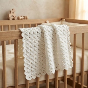 May include: A white crocheted baby blanket with a textured diamond pattern and a scalloped edge. The blanket is draped over the wooden railing of a crib, showcasing the intricate yarn work and the soft, off-white color of the textile material.