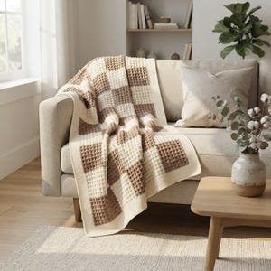 Peut inclure: Un plaid en maille gaufrée présentant un motif à grands carreaux alternant des tons crème et marron clair. La couverture possède une surface texturée en forme de grille et est drapée sur l'accoudoir et l'assise d'un canapé de couleur neutre.