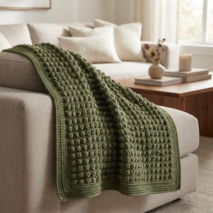 Puede incluir: Una manta de ganchillo color verde salvia con un patrón de puntos de burbuja texturizados en todo el cuerpo principal y un borde plano y sólido. La manta está colocada sobre el brazo de un sofá de color neutro, resaltando los nudos de hilo.