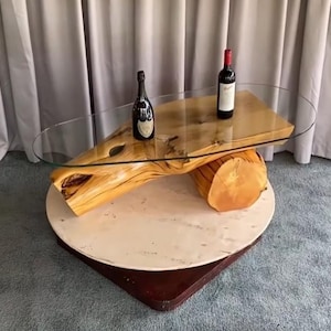 Peut inclure: Une table basse dotée d'un plateau ovale en verre transparent reposant sur deux bûches de bois clair naturel. La table est posée sur une base ronde blanche et une plateforme carrée sombre. Deux bouteilles de vin sont posées sur le verre.