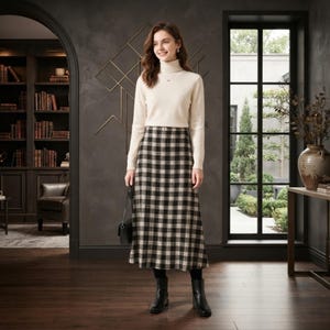 Puede incluir: Un suéter de cuello alto color crema de manga larga combinado con una falda midi a cuadros negros, blancos y beige. El conjunto incluye botines negros y un pequeño bolso de mano negro que se sostiene con una mano.