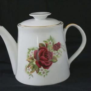 Può includere: Una teiera in porcellana bianca con un motivo floreale raffigurante una grande rosa rosso scuro, boccioli di rosa più piccoli e minuscoli fiori bianchi con foglie verdi. Il coperchio abbinato è rifinito con un bordo dorato.